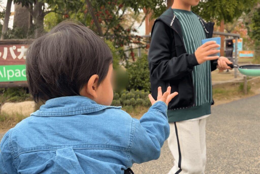 ラクダの前で、次男が長男に「えさちょうだい」と手を伸ばしている様子